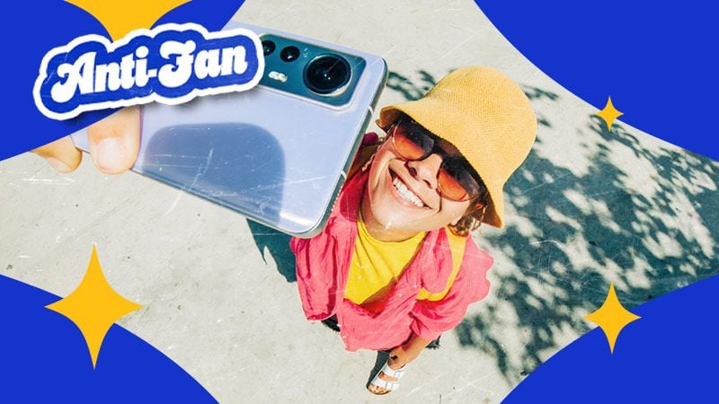 Smiling person in a bright yellow hat and pink jacket takes a selfie with a phone. Blue and gold star graphics and "Anti-Fan" text overlay. Cheerful mood.