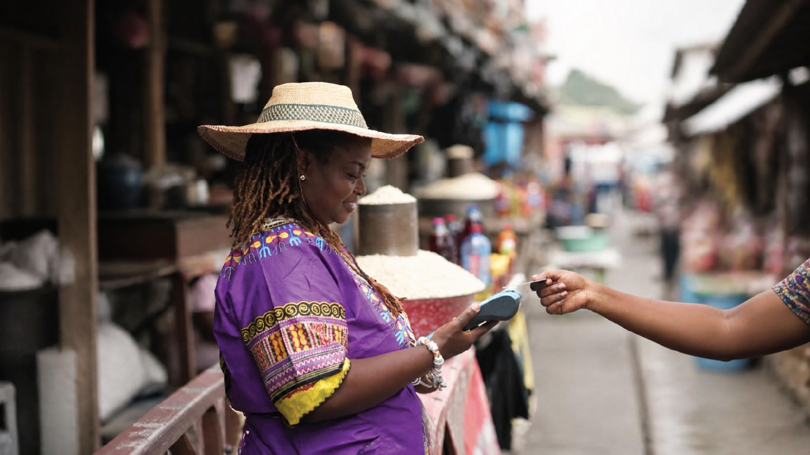 A woman in a vibrant purple dress and straw hat uses a card reader for payment at a market stall.