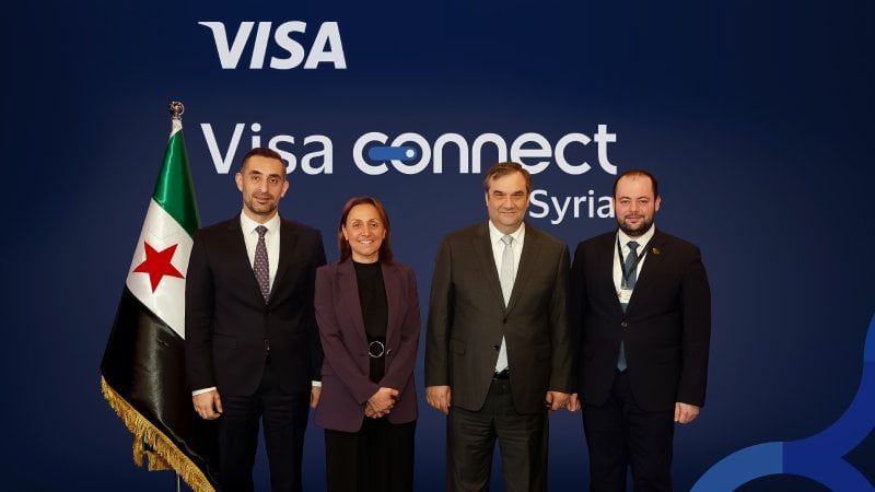 Four individuals in formal attire stand in front of a "Visa Connect Syria" backdrop. A flag with red stars is positioned to their left, creating a professional and diplomatic atmosphere.