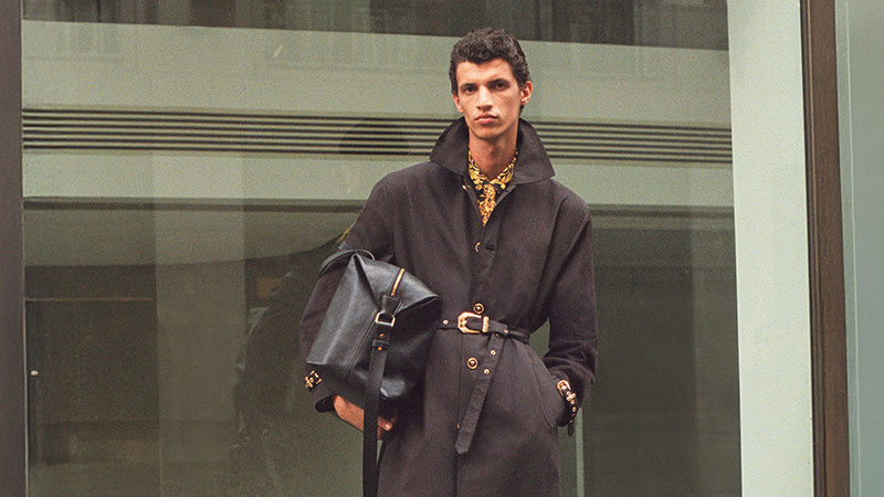 Young man stands confidently in front of glass building, wearing a dark trench coat with a patterned shirt. He holds a large black bag. Fashionable and poised.