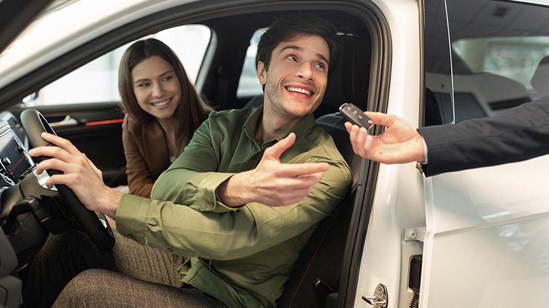 A smiling man in a car receives keys from a person outside, with a woman beside him