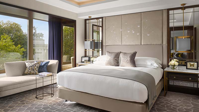 Luxurious bedroom with a large bed, plush pillows, and elegant decor. A cozy sofa by a window overlooks greenery, creating a serene atmosphere.