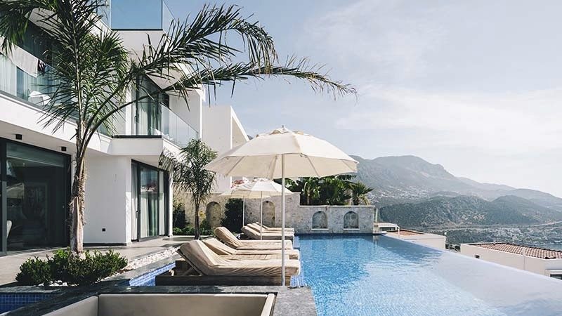 Luxurious poolside scene with lounge chairs under umbrellas by an infinity pool, overlooking scenic mountains and a coastal view