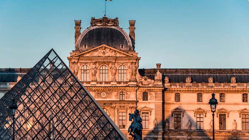 The image shows the Louvre Museum with its iconic glass pyramid in the foreground at sunset. The baroque architecture is bathed in warm light.