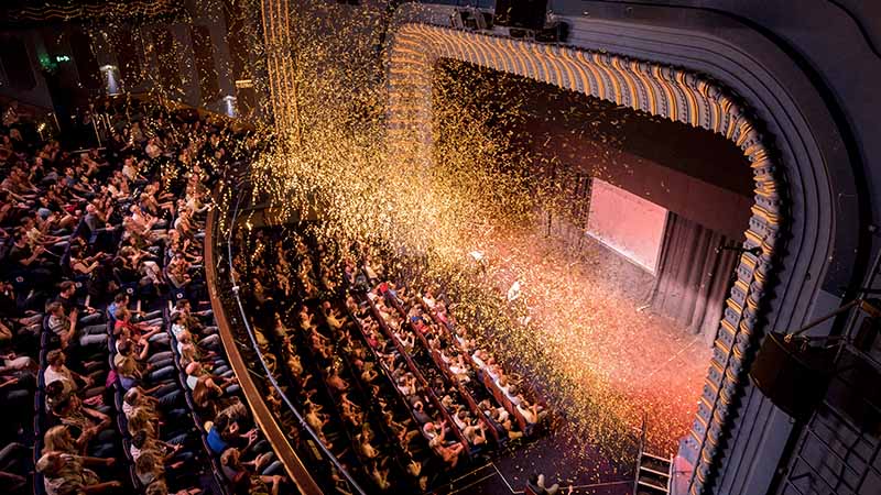 A theater filled with an audience watching a stage show, as golden confetti falls from above, creating a festive and celebratory atmosphere.