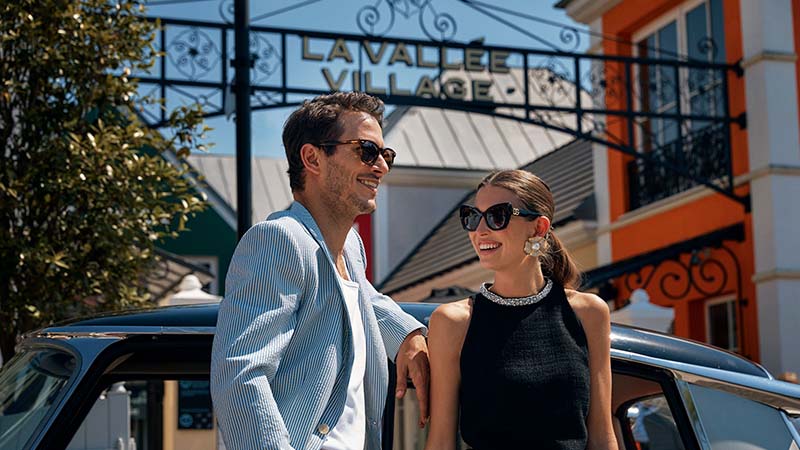 A stylish couple in sunglasses smiles while leaning against a vintage car at La Vallée Village, an upscale shopping venue, under a sunny sky.