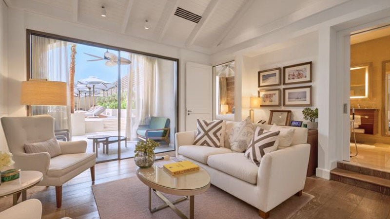 Cozy living room with a white couch, patterned cushions, and an armchair. A sliding glass door opens to a sunny patio. Warm lighting adds a welcoming feel.