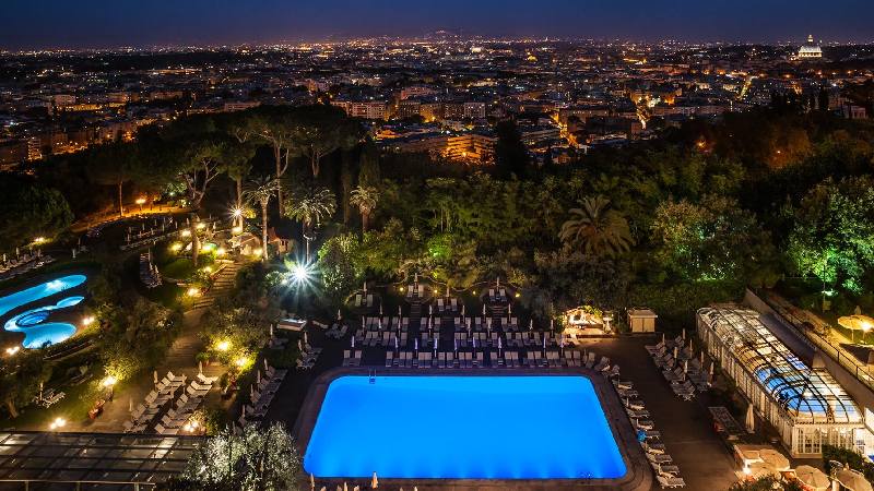 Nighttime aerial view of a resort pool and gardens overlooking a glowing city skyline