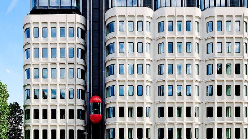 Modern high-rise building facade with rounded windows and a red external elevator