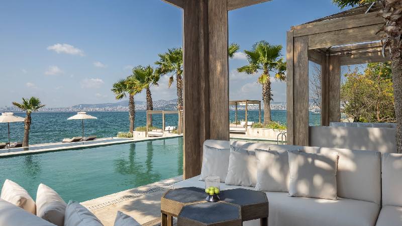 Seaside poolside lounge with white cushions, palm trees, and panoramic ocean views