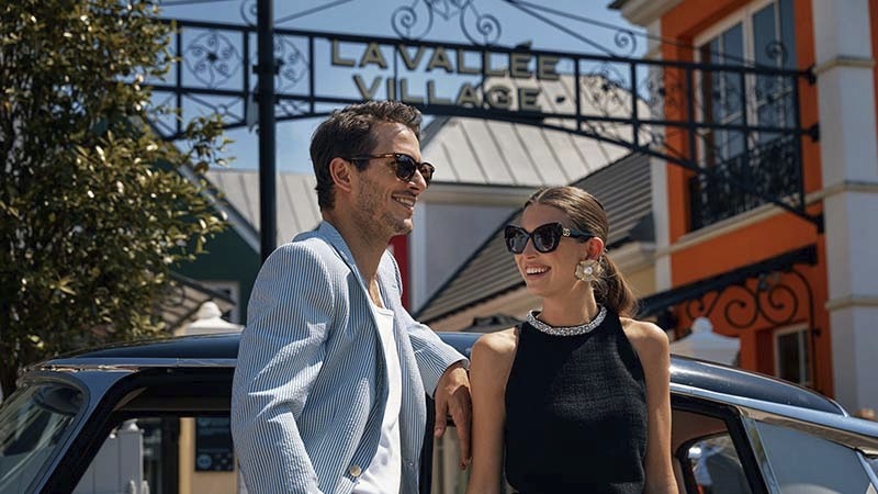 Stylish couple wearing sunglasses standing by a car outside La Vallée Village