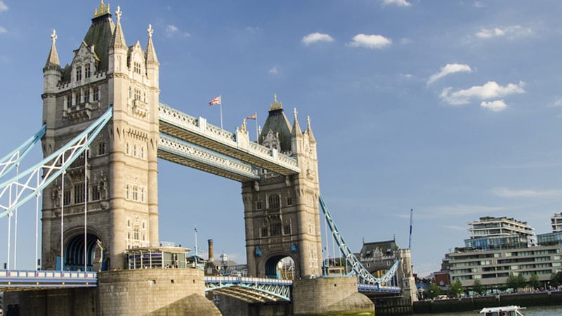 Tower Bridge in London under a clear blue sky. The iconic bridge, with its two large towers and blue suspension cables, spans the River Thames.