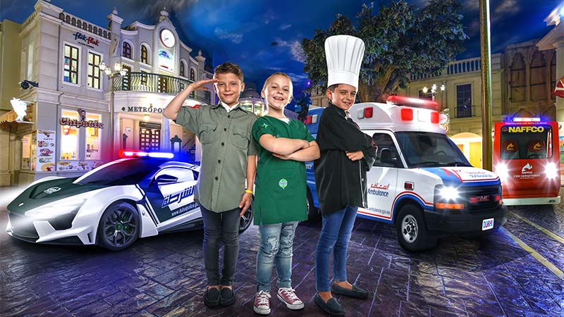 Three children in uniforms stand in front of a police car, ambulance, and fire truck on a city street, with buildings in the background at dusk.