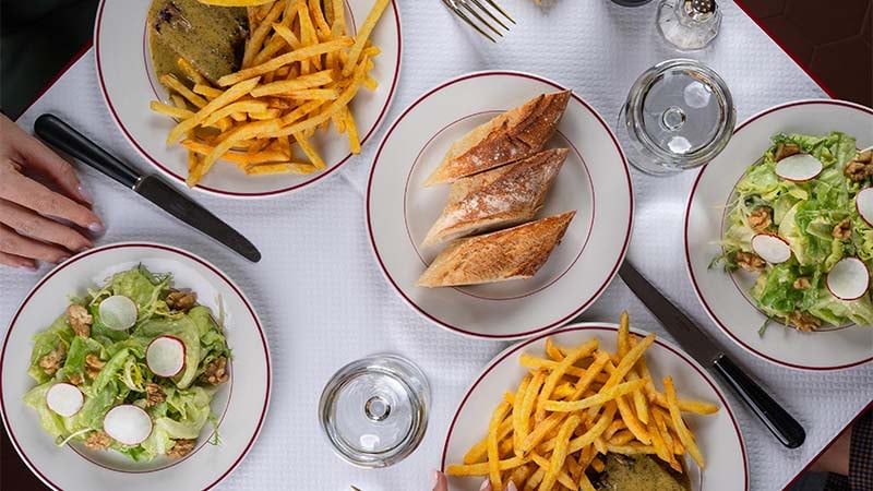 Four plates with fries, salad with radishes and walnuts, and bread are arranged on a white tablecloth with water, cutlery, and a hand visible on the left.