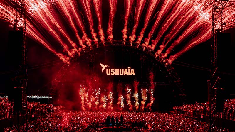 A large crowd at the Ushuaïa stage watches red fireworks and lights arching over the stage during a nighttime concert or event.
